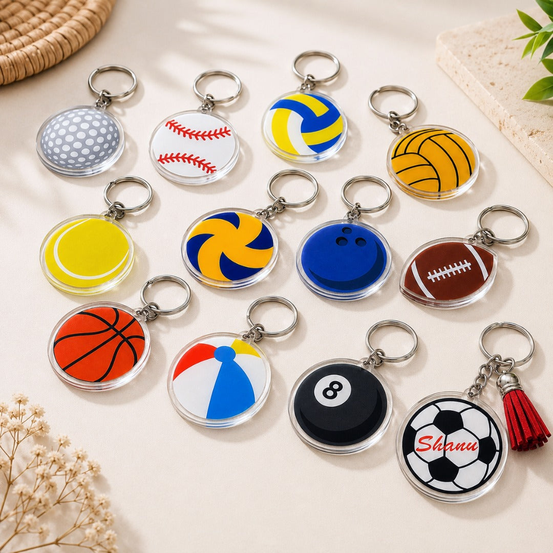 Twelve round acrylic sports ball keychains displayed on a light beige surface, featuring golf, baseball, volleyball, tennis, basketball, bowling, American football, soccer, beach ball, billiard 8-ball, and personalized soccer ball SVG, PNG, DXF, and PDF designs for Cricut, laser cutting, and DIY keychain projects.